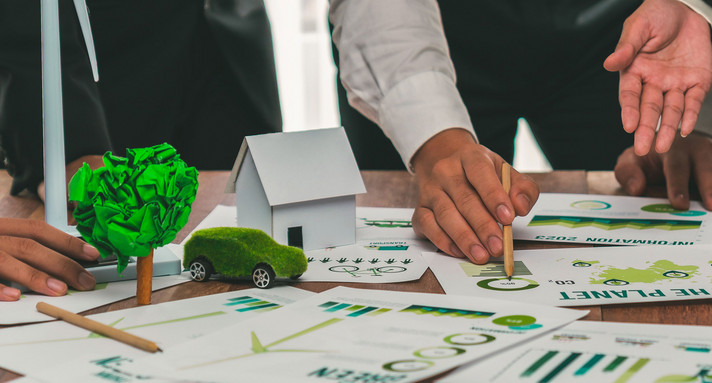 Menschen sind über einen Tisch gebeugt auf als Figuren ein mit Gras überwachsenes Auto und ein Haus aus Papier stehen.