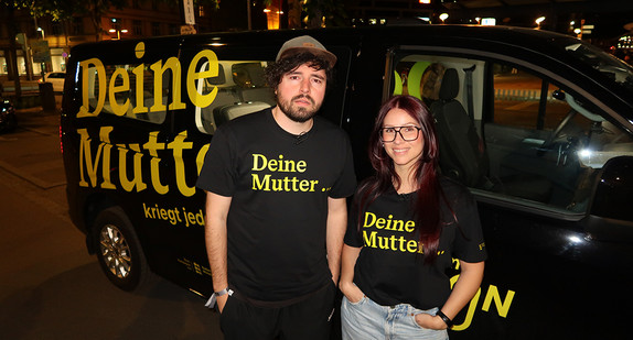Ein Mann mit Cap und eine Frau stehen vor einem Auto mit der Aufschrift DEINE MUTTER und haben schwarze T-Shirts an mit gelber Schrift "Deine Mutter..."